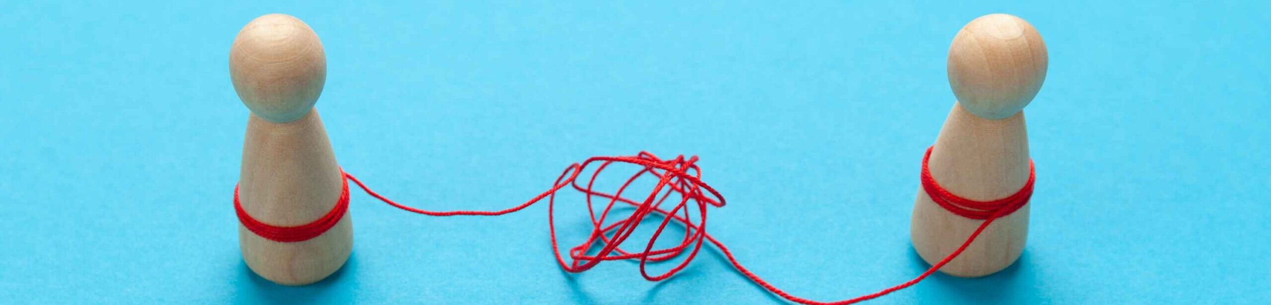 Zwei Holzfiguren, verbunden durch einen verknoteten roten Faden – Symbol für zwischenmenschliche Verstrickungen und emotionale Blockaden.