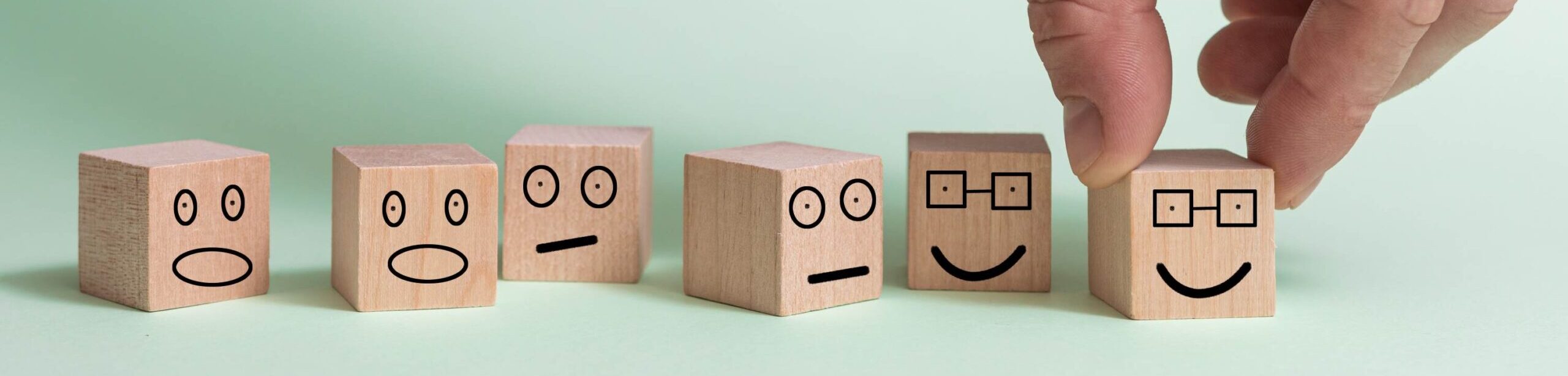 Holzwürfel mit verschiedenen Gesichtsausdrücken, eine Hand wählt einen lachenden Würfel mit Brille – symbolisiert den bewussten Umgang mit Persönlichkeitsanteilen.
