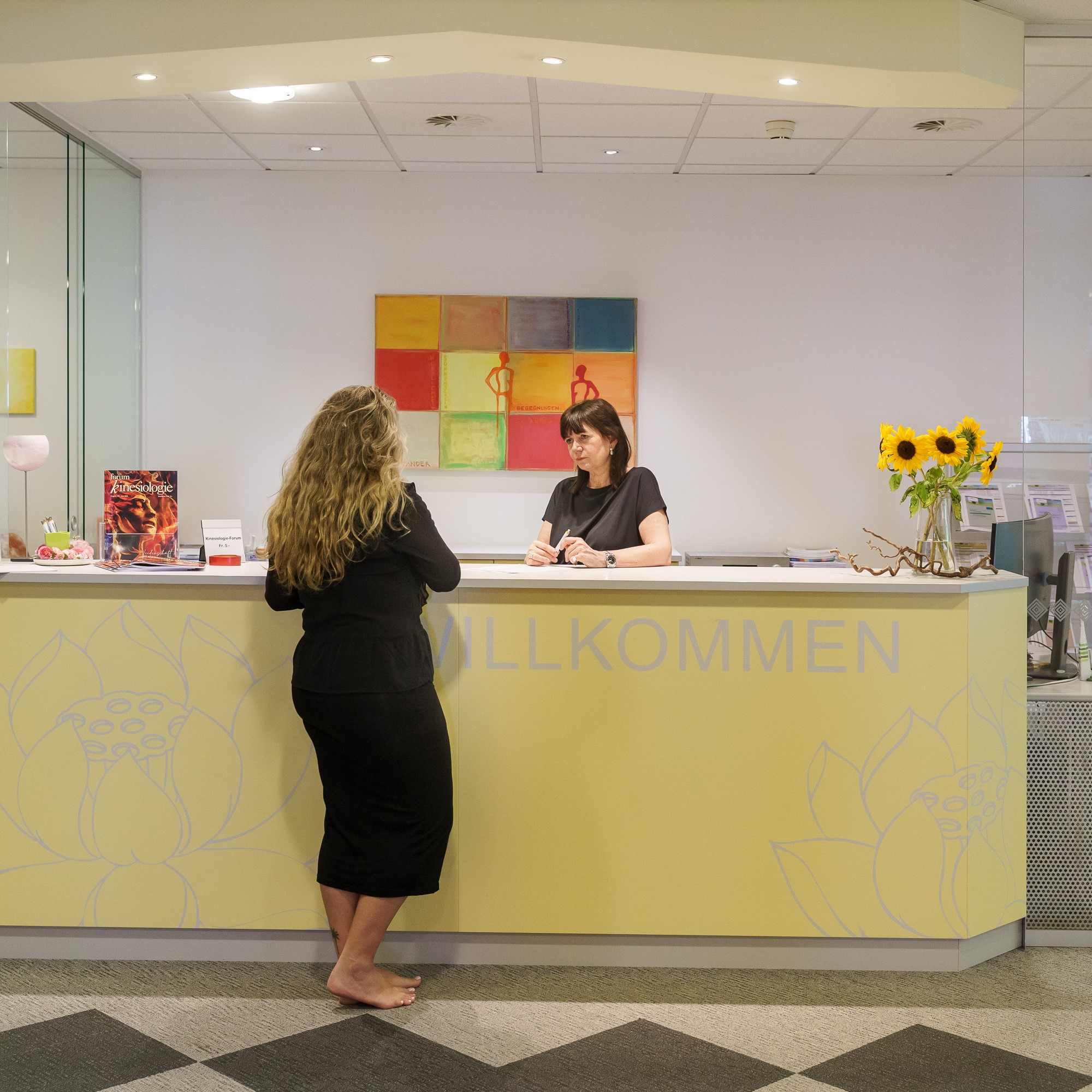 Zwei Frauen stehen im Gespräch am Empfangstresen der APAMED mit der Aufschrift „Willkommen“.