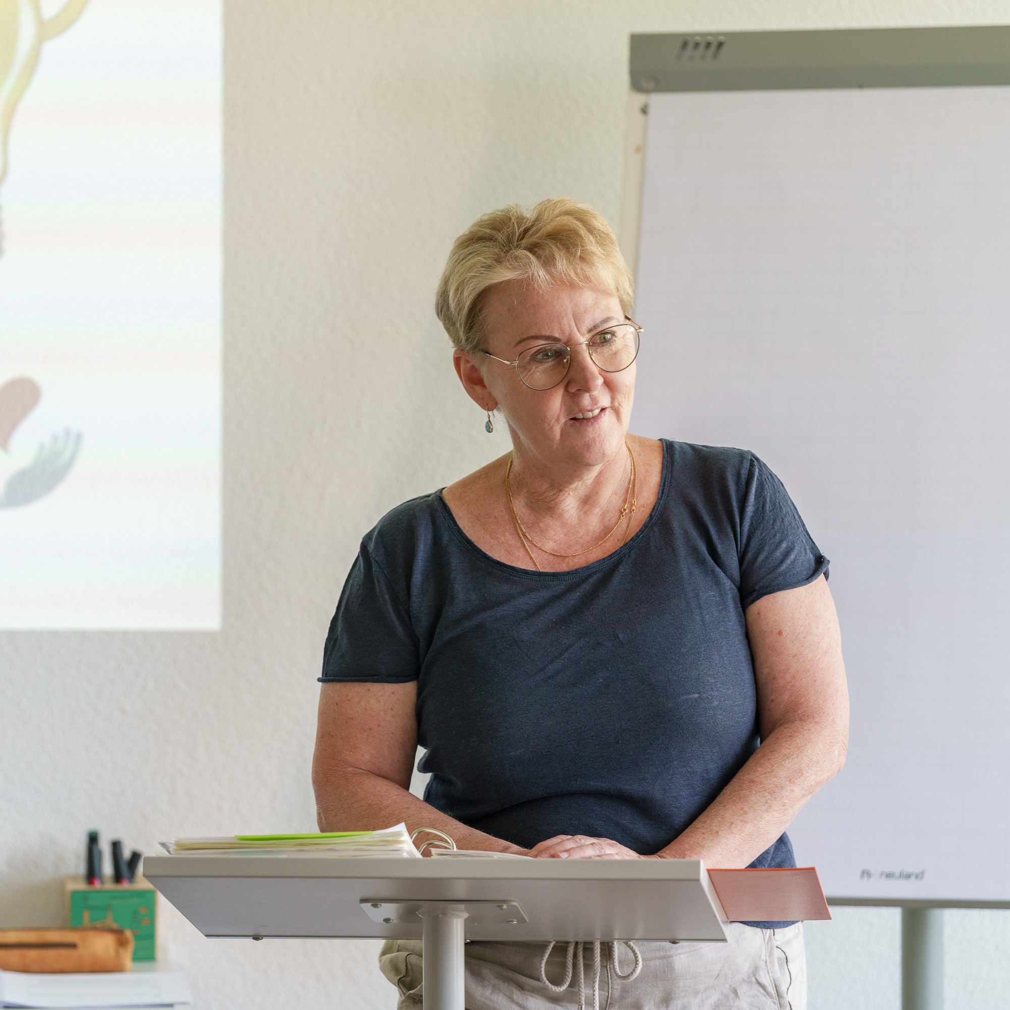 APAMED-Dozentin steht am Pult und spricht im Unterricht vor einer Präsentationsfolie und einem Flipchart.