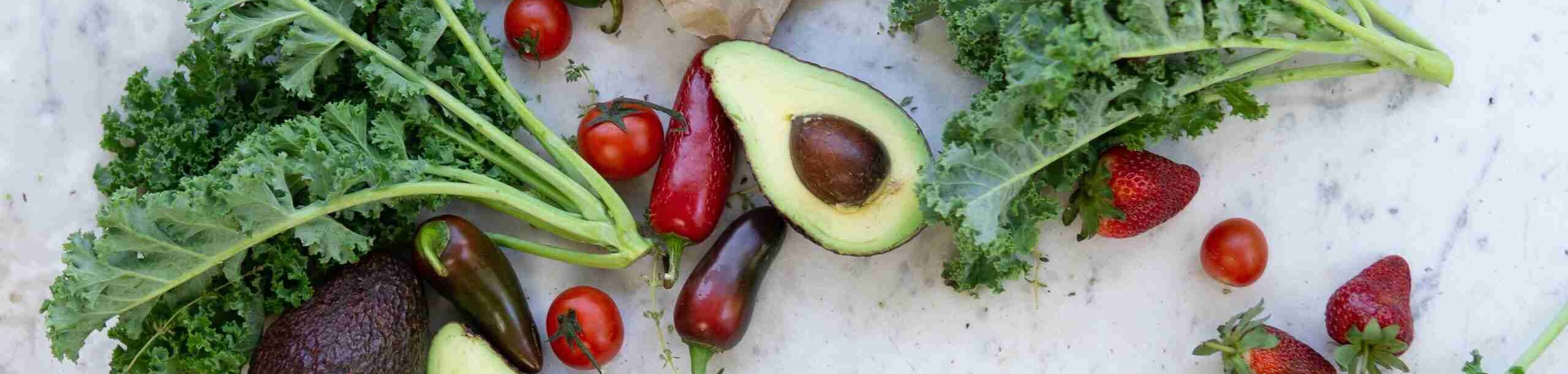 Ausbildung Ernährungsberatung: Frisches Gemüse und Obst wie Grünkohl, Avocado, Cherrytomaten, Peperoni und Erdbeeren liegen auf einer hellen Steinplatte.