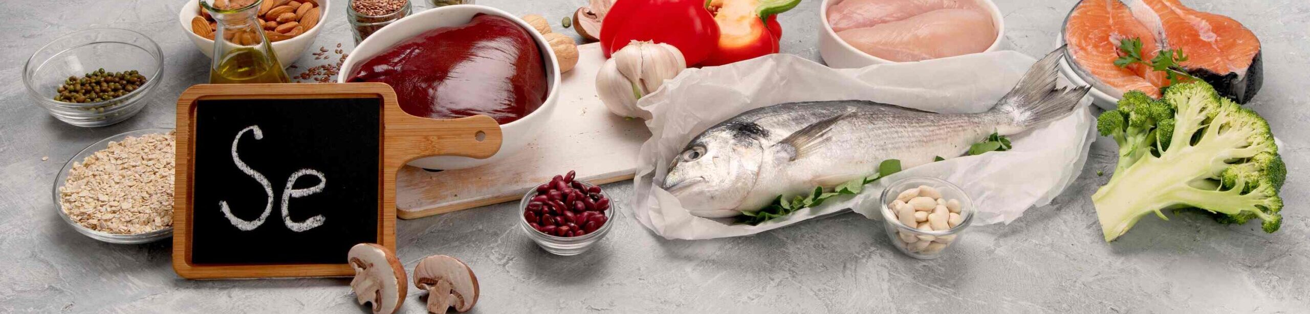 Lebensmittel mit hohem Selengehalt wie Fisch, Leber, Nüsse und Hülsenfrüchte auf grauem Untergrund, Schild mit „Se“ im Vordergrund