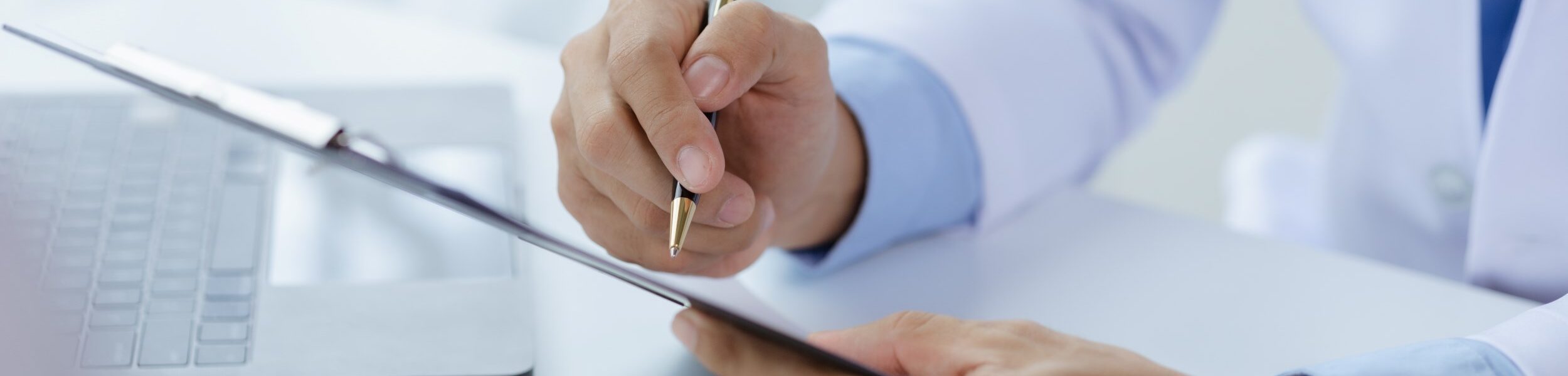 Nahaufnahme eines Arztes oder Therapeuten im Gespräch – er zeigt mit einem Stift auf ein Klemmbrett mit Unterlagen.