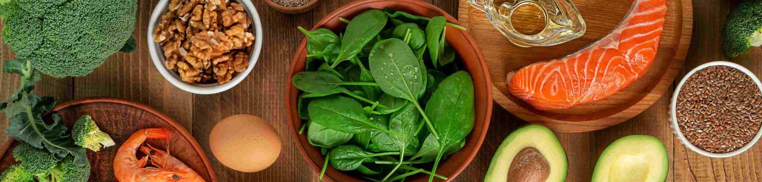 Ausgewogene Lebensmittel wie Fisch, Avocado, Nüsse, Eier und Gemüse auf einem Holztisch – symbolisch für Proteine, Fette und Kohlenhydrate