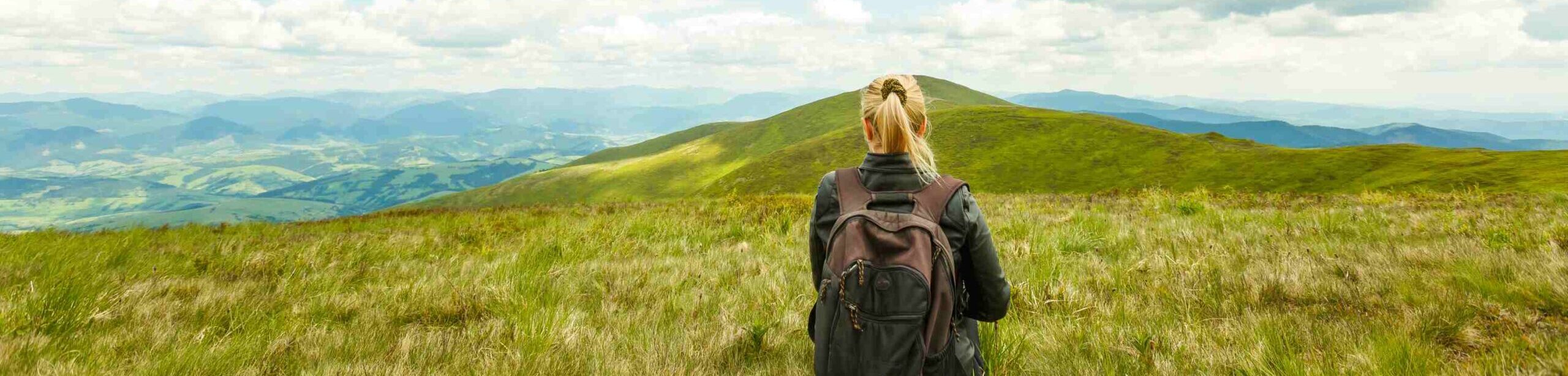 Frau mit Rucksack sitzt entspannt auf einer grünen Bergwiese mit Blick auf weite Hügelketten – Symbol für Stressabbau und mentale Klarheit