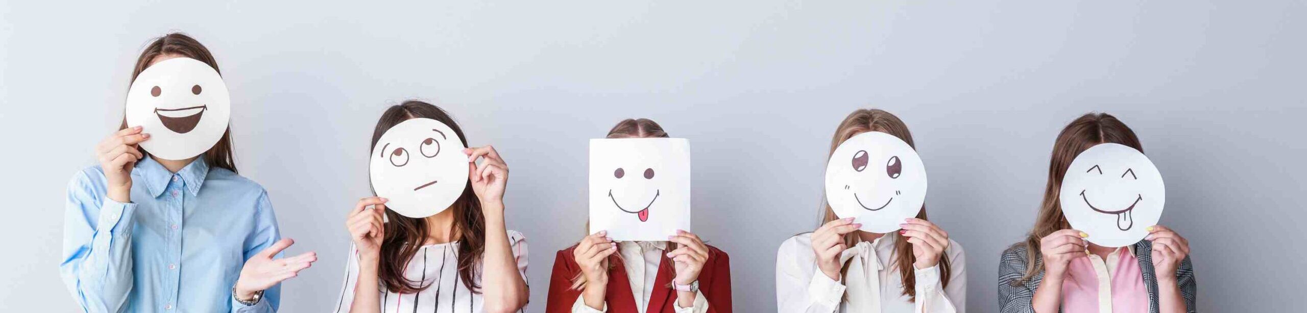 Fünf Frauen halten gezeichnete Gesichtsausdrücke vor ihr Gesicht, die verschiedene Emotionen darstellen – von Freude bis Unsicherheit.