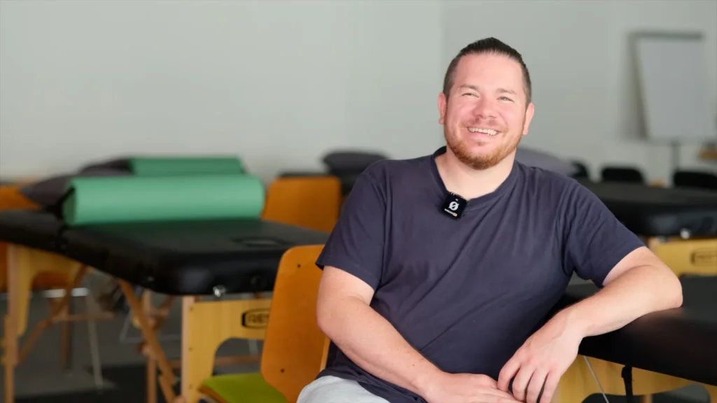 Martin, Absolvent und Dozent der Kinesiologie Ausbildung, sitzt lächelnd im Unterrichtsraum der APAMED Fachschule.